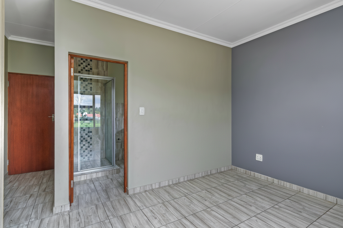 A bright bedroom featuring wood-look tiled flooring, a neutral and grey accent wall, and an en-suite bathroom with a glass-enclosed shower.