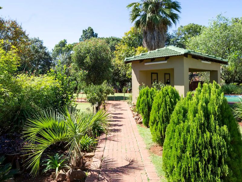 A well-maintained garden featuring a brick-paved pathway leading to a covered gazebo, surrounded by mature trees and manicured shrubs.