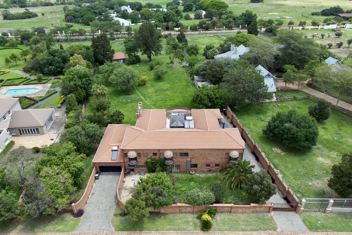 Aerial view of a large brick residence with a tiled roof, featuring a paved driveway, perimeter wall, and expansive surrounding garden grounds.