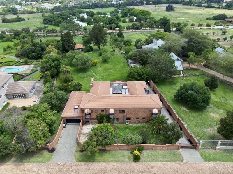 Aerial view of a large brick residence with a tiled roof, featuring a paved driveway, perimeter wall, and expansive surrounding garden grounds.
