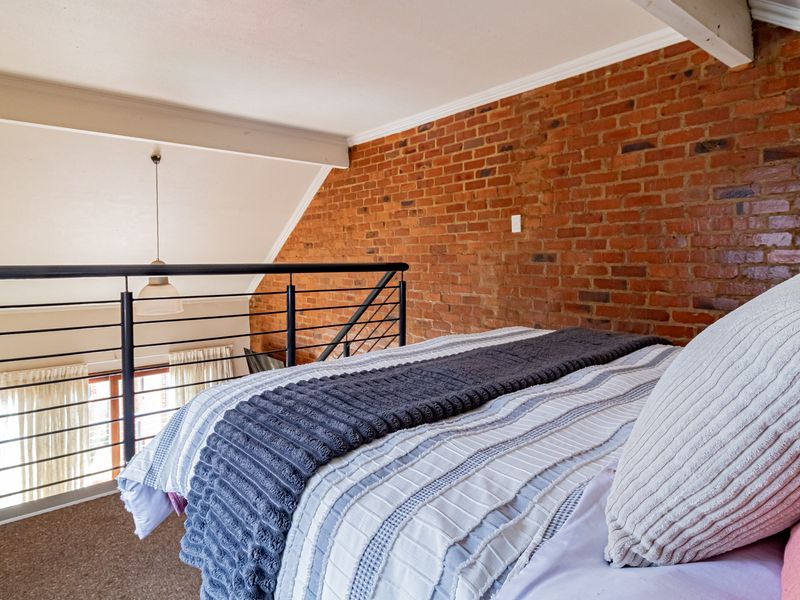 A loft-style bedroom featuring an exposed brick feature wall, vaulted ceiling with exposed beams, and a metal balustrade overlooking the lower level.