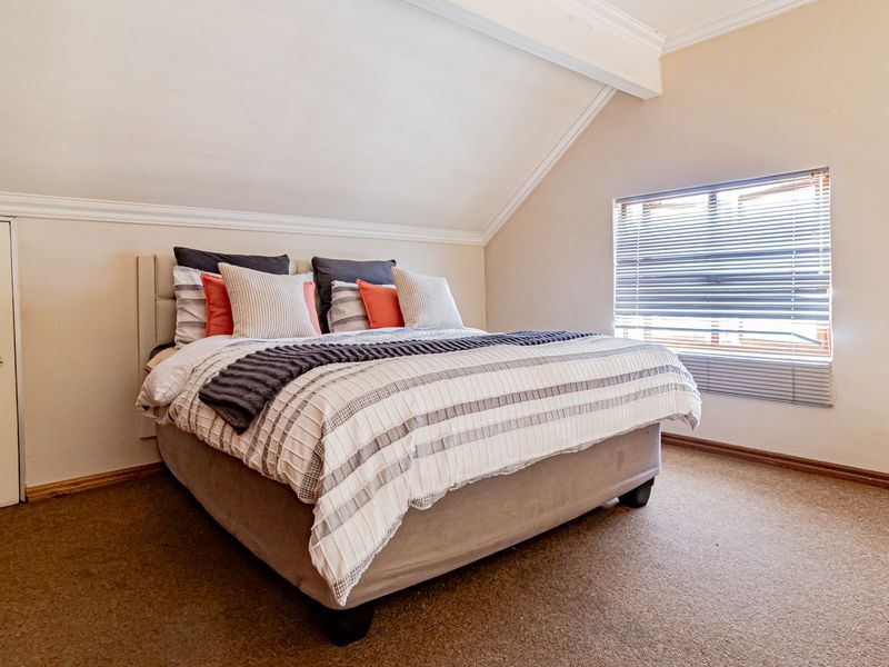 A cozy bedroom featuring a sloped ceiling, built-in storage, and carpeted flooring, illuminated by natural light through a window with venetian blinds.