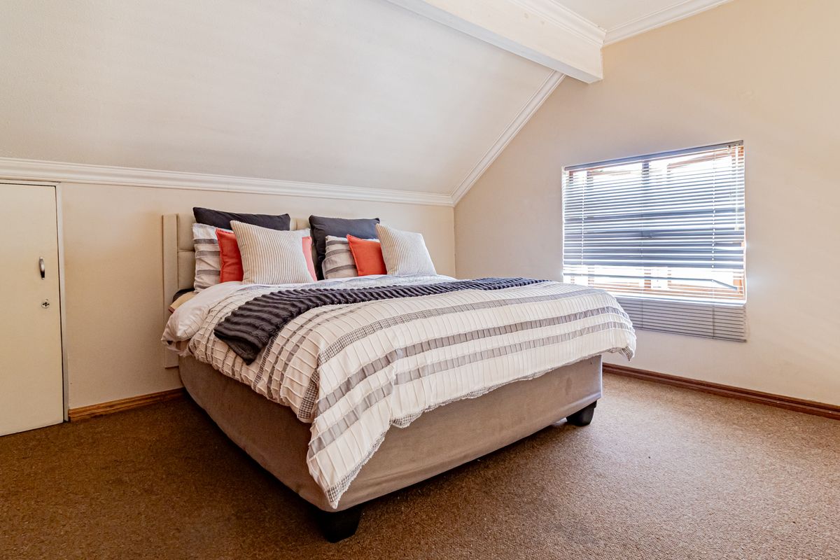 A cozy bedroom featuring a sloped ceiling, built-in storage, and carpeted flooring, illuminated by natural light through a window with venetian blinds.