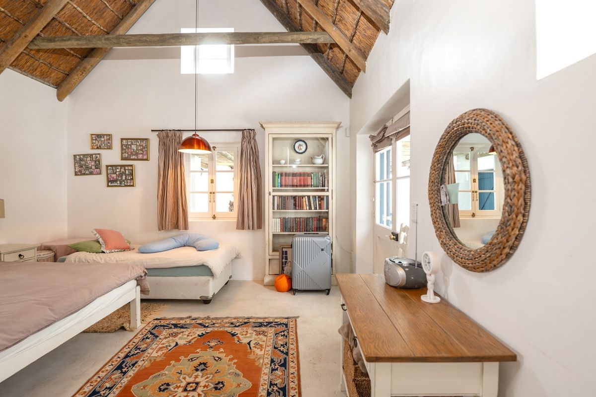 Spacious bedroom featuring a high vaulted ceiling with exposed timber rafters, thatched roofing, built-in shelving, and natural light through casement windows and french doors.