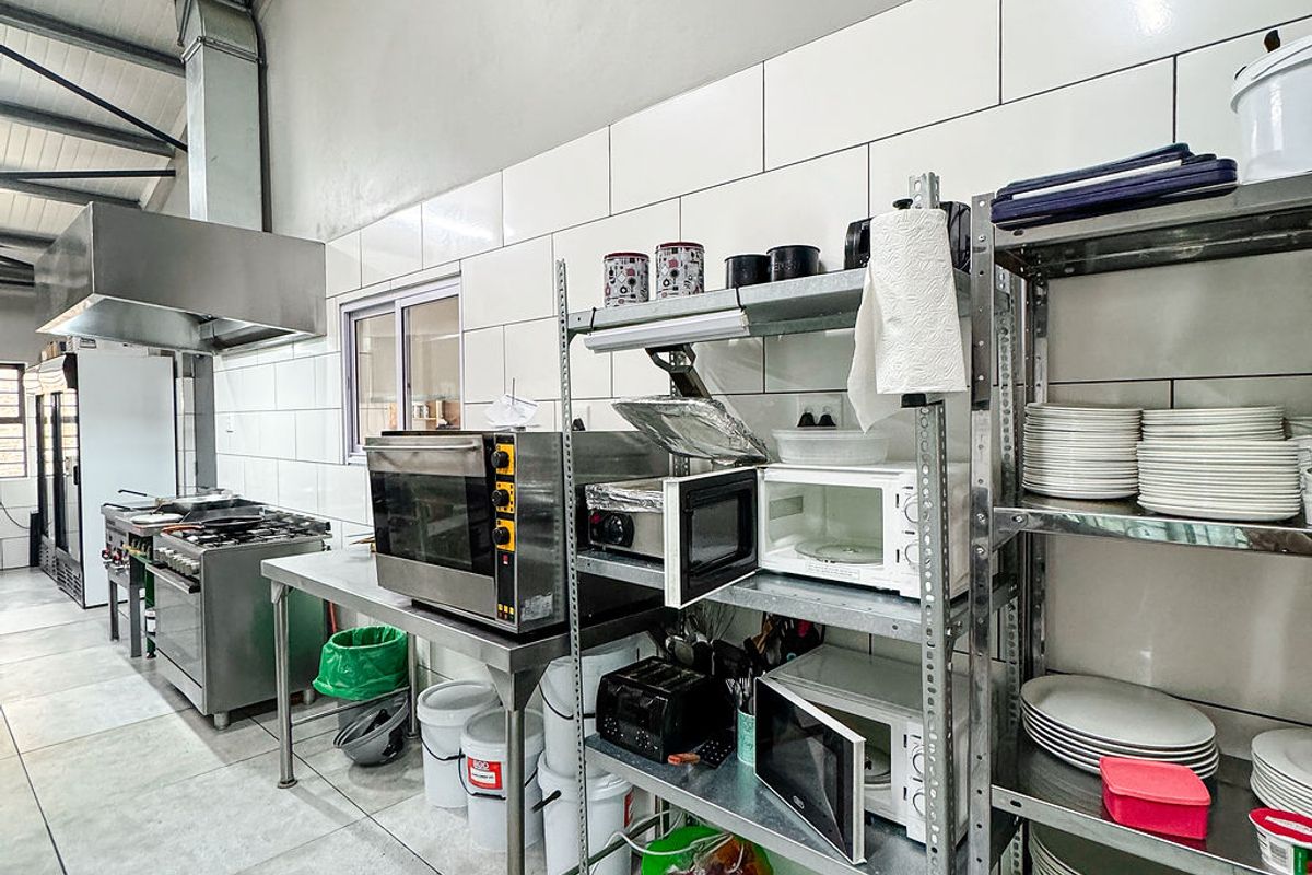 A functional commercial-style kitchen featuring stainless steel appliances, industrial extractor hood, tiled surfaces, and exposed roof trusses for a spacious, utilitarian workspace.