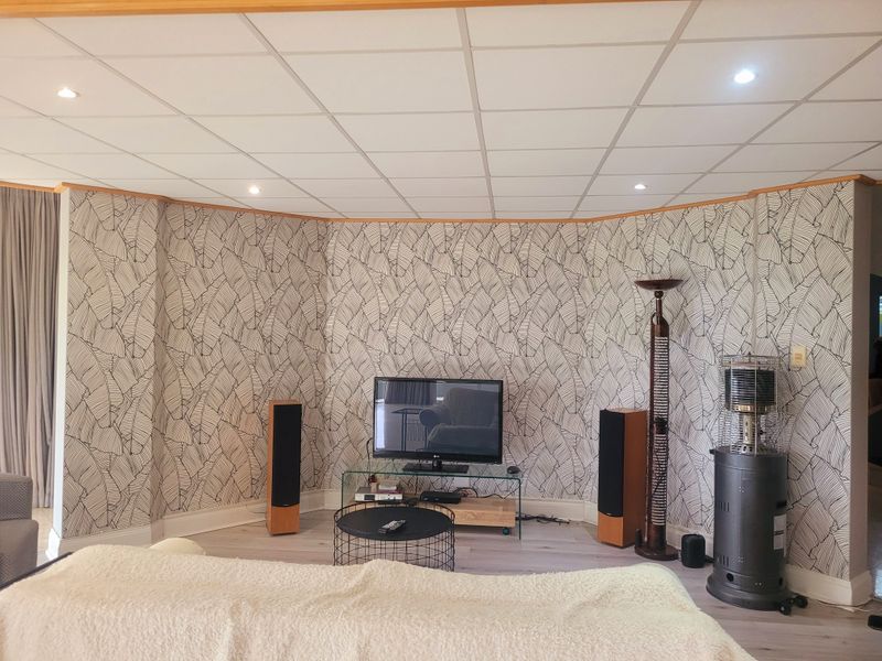 A living room featuring a curved wall with patterned wallpaper, recessed downlights, a suspended ceiling, and light-toned laminate flooring.