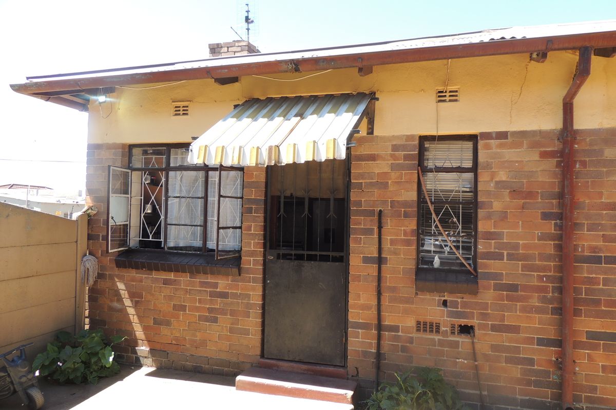 Rear exterior view of a brick house featuring a metal awning, security gate, and steel-framed windows with security bars.