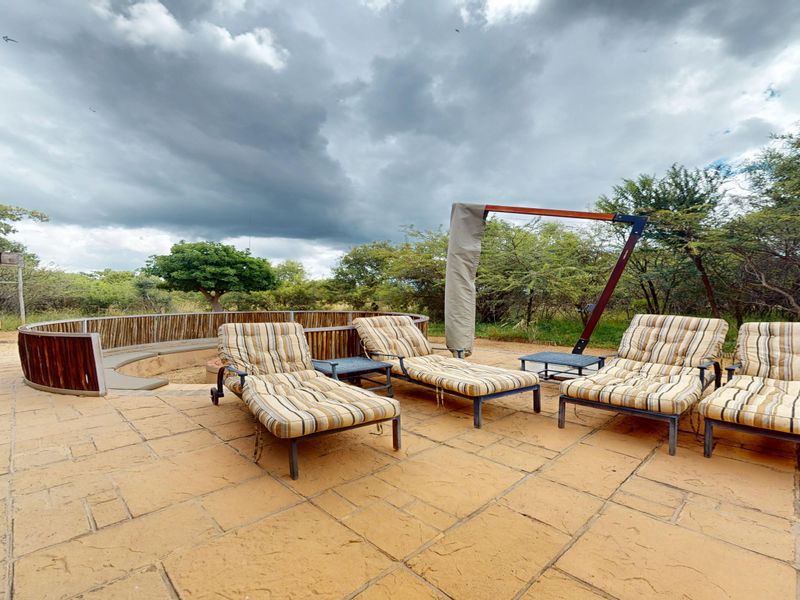 An outdoor patio area featuring paved flooring, a curved wooden retaining wall, and a sunken fire pit, set against a natural bushveld backdrop.