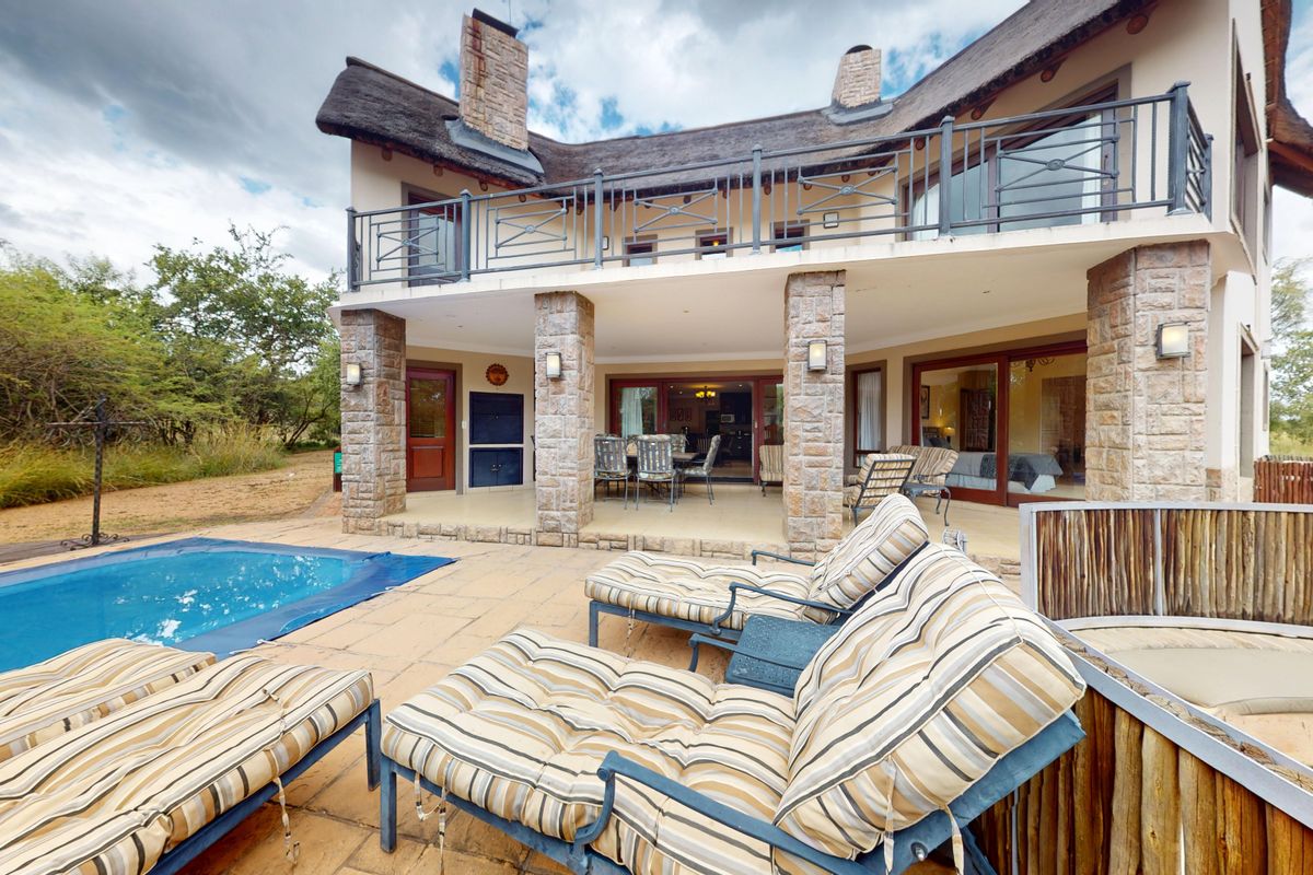 Rear view of a double-story house featuring a swimming pool, covered patio, balcony with metal railings, and stone-clad pillars under a thatched roof.