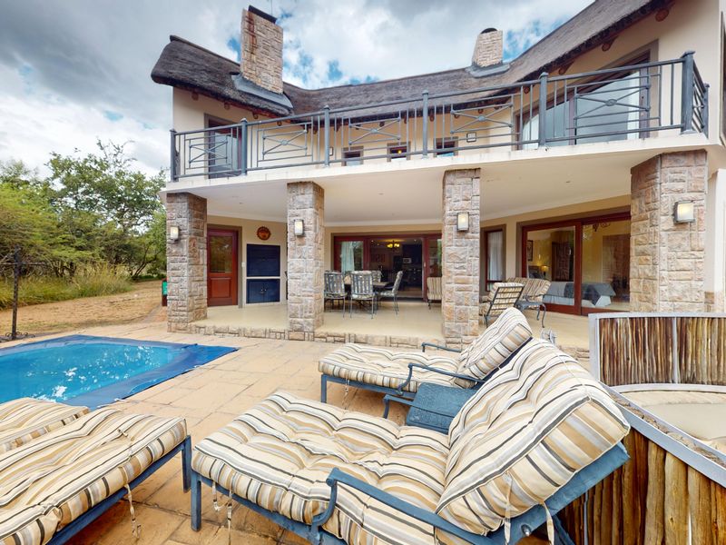 Rear view of a double-story house featuring a swimming pool, covered patio, balcony with metal railings, and stone-clad pillars under a thatched roof.