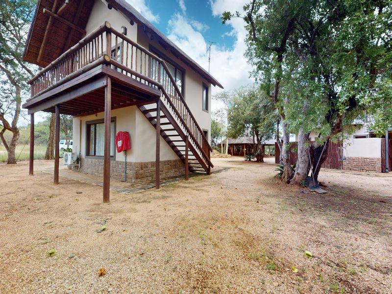 A two-story house featuring a thatched roof, wooden balcony, external staircase, and stone cladding, set within a natural bushveld environment.