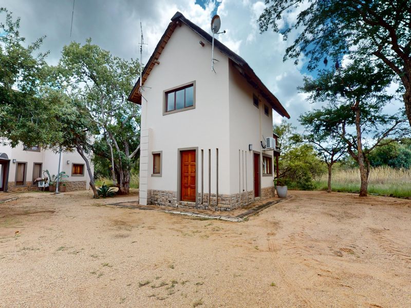 A two-story house featuring a thatched roof, stone cladding base, and timber entrance door, situated in a natural, wooded setting.