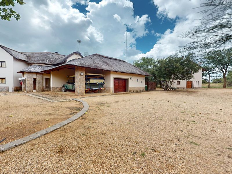 A spacious property exterior featuring a traditional thatch roof, stone facade detailing, a covered carport, and a large gravel driveway.
