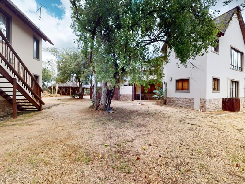 Rear view of the property featuring stone-clad exterior walls, an external timber staircase, and a spacious gravel courtyard area with mature trees.