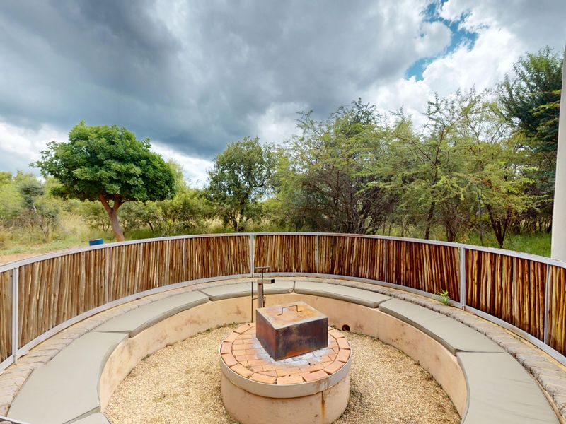 A sunken outdoor fire pit area featuring built-in circular seating and a brick fire pit, surrounded by natural timber fencing in a bushveld setting.