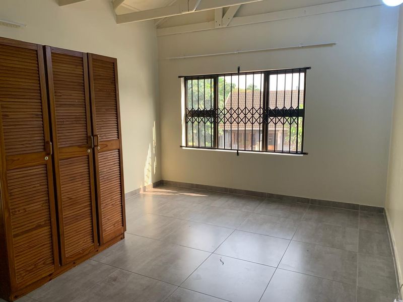 A bright, empty bedroom featuring tiled flooring, exposed ceiling rafters, and a built-in wooden wardrobe with security bars on the window.