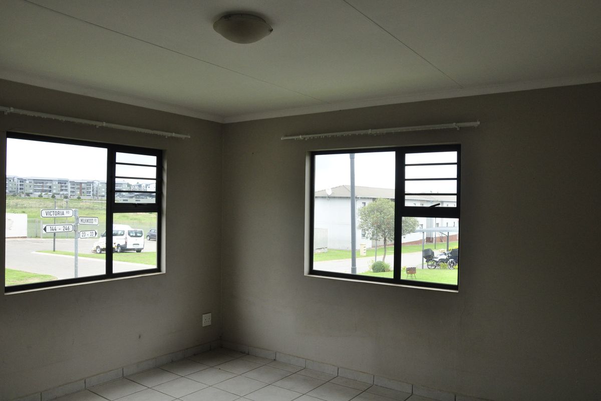 A bright, empty bedroom featuring neutral wall tones, tiled flooring, and large windows providing natural light and views of the surrounding complex.