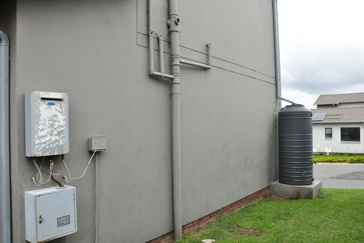 Exterior side view of the townhouse featuring a wall-mounted gas geyser, external plumbing, and a rainwater collection tank.
