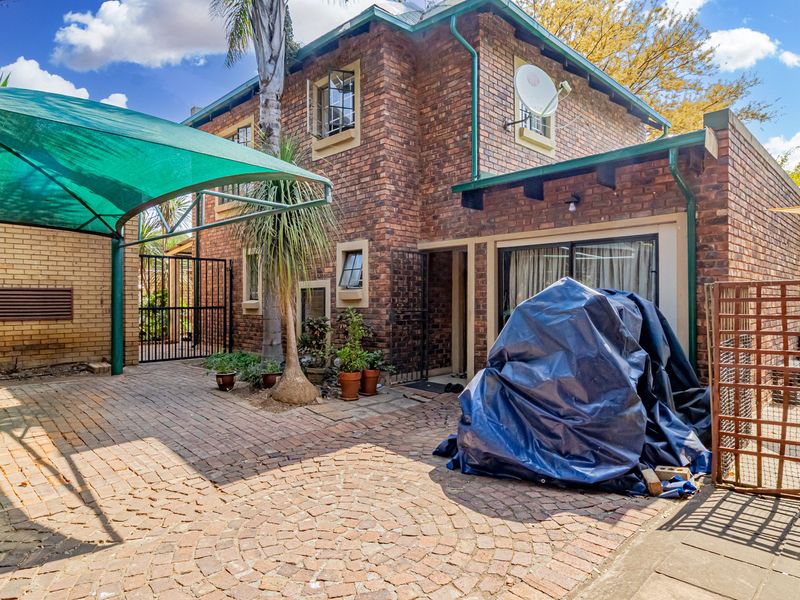 Front exterior view of a face brick townhouse featuring a paved driveway, shade cloth carport, and secure gated entrance.