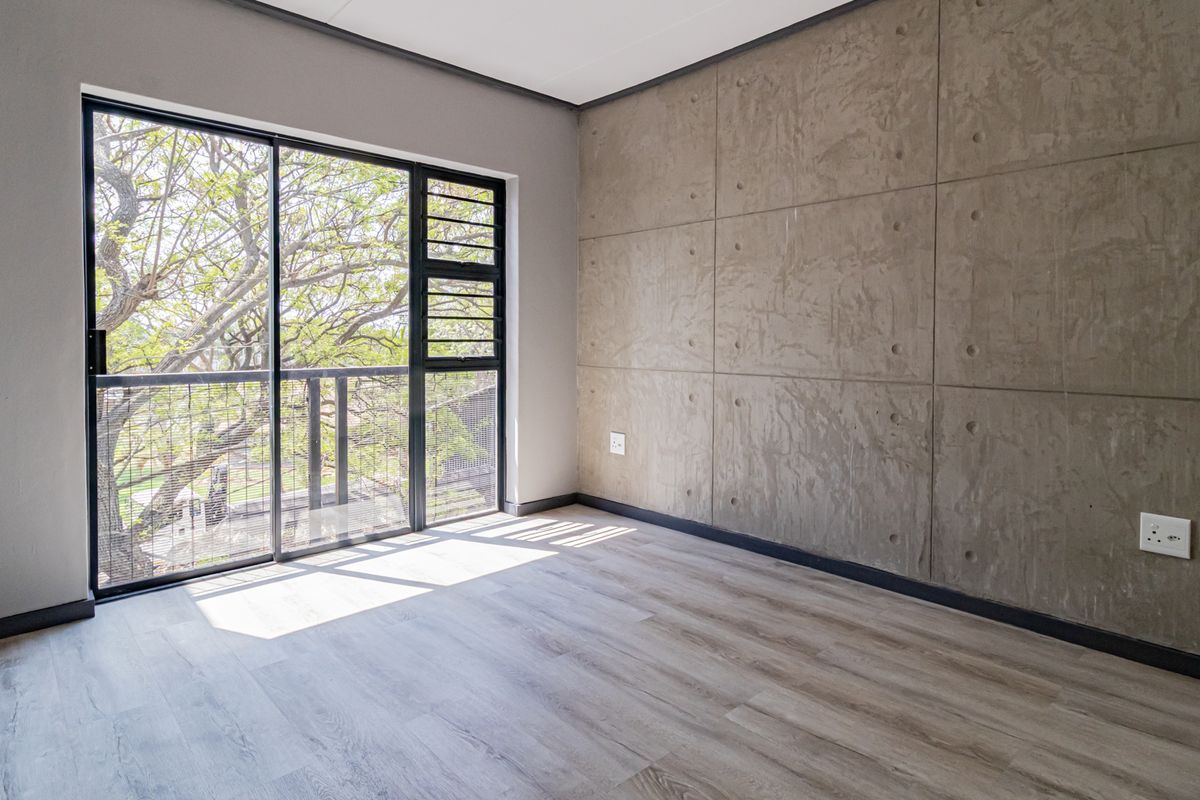A bright, modern bedroom featuring a raw concrete accent wall, light wood-look laminate flooring, and a large sliding glass door with balcony access.