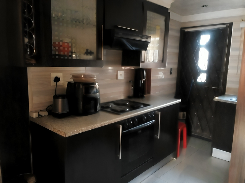 A compact kitchen featuring dark cabinetry, a built-in oven with electric hob, extractor fan, and tiled backsplash, leading to an exterior door.
