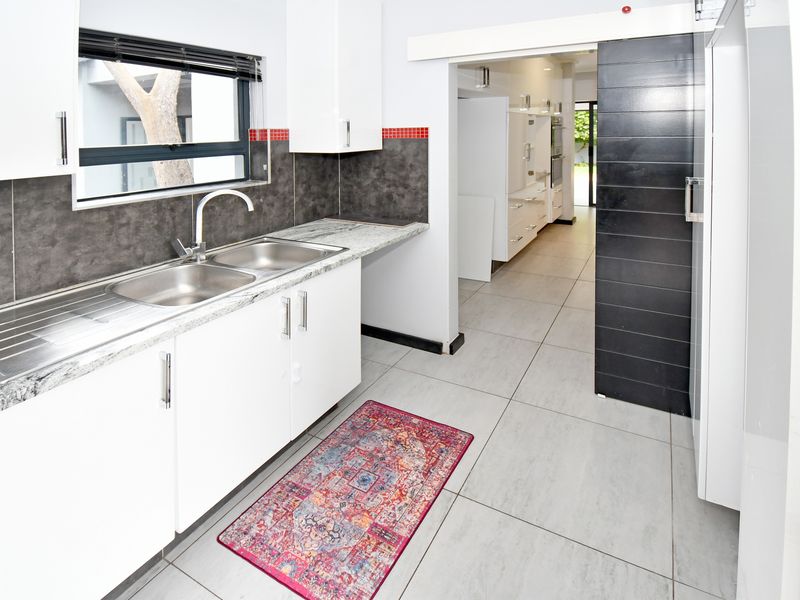A clean, modern scullery featuring white cabinetry, granite countertops, a double sink, and large format floor tiles, leading into the main kitchen area.