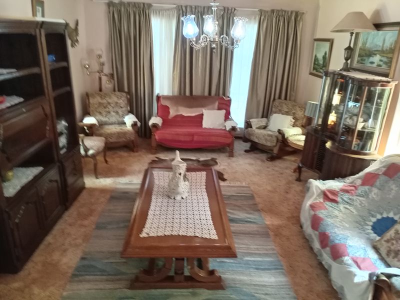 A traditional living room featuring a central chandelier and large window curtains, currently furnished with dated decor and carpeting.