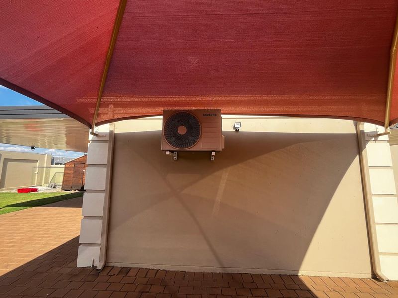 A covered patio area featuring a shade sail and a wall-mounted air conditioning condenser unit, overlooking a paved outdoor space.