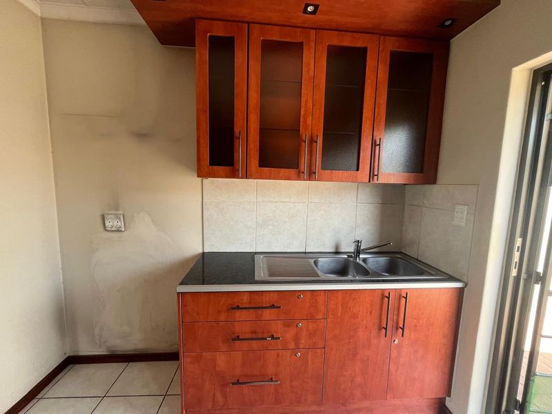 A compact kitchen area featuring wooden cabinetry, a stainless steel double sink, and tiled backsplash, currently requiring wall repairs and cosmetic updates.