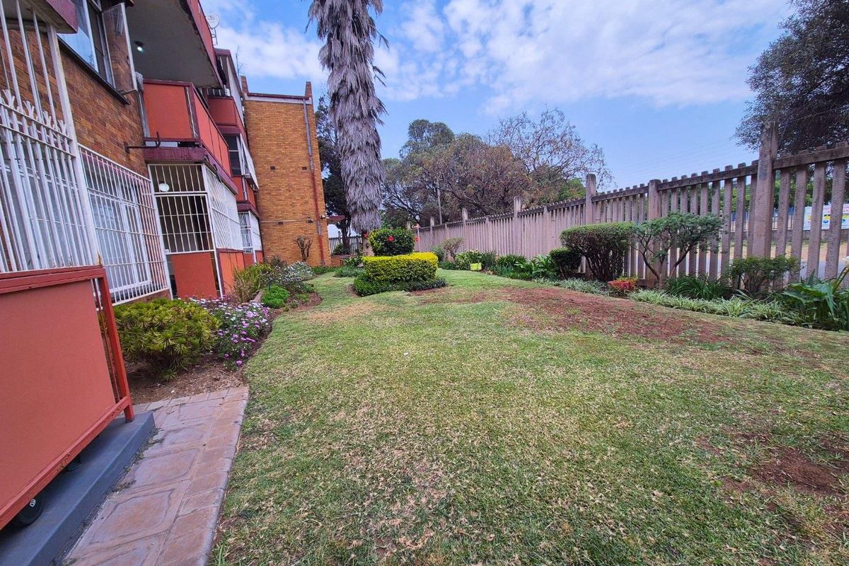 A view of the apartment building's rear garden area, featuring a lawn, established shrubs, and a perimeter concrete fence.