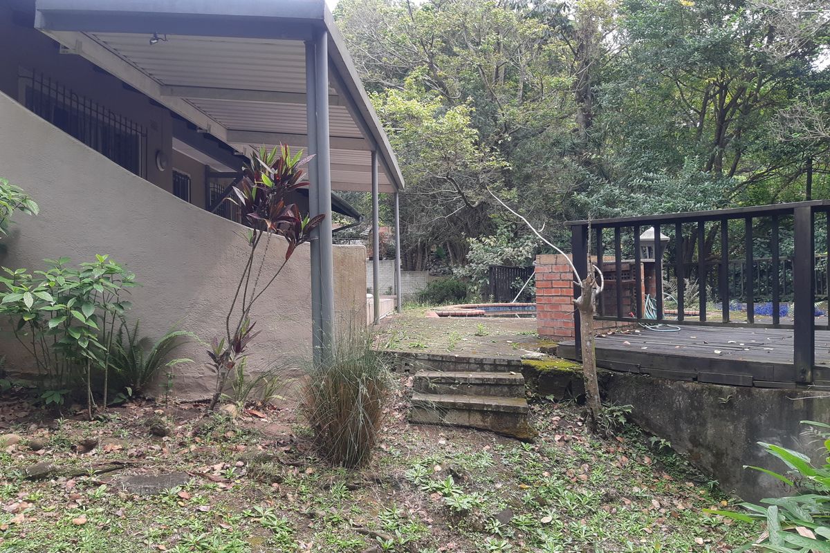 Rear exterior view featuring a covered patio, timber decking, and a brick built-in braai area set within a mature, overgrown garden.