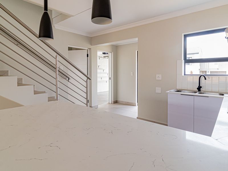 Modern open-plan kitchen featuring a stone countertop, gas hob, and sleek staircase with stainless steel railing in a bright, neutral interior.