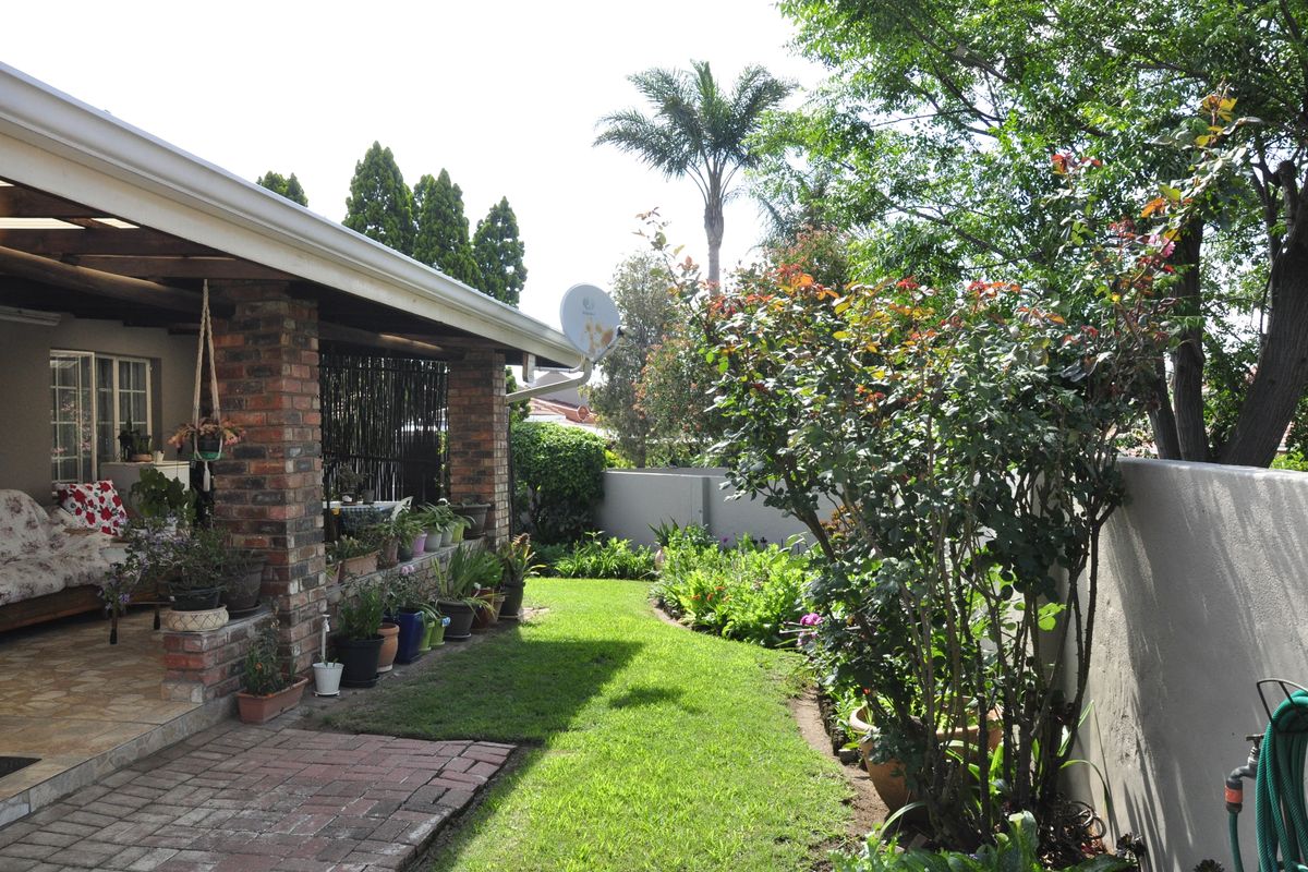 A well-maintained rear garden featuring a covered brick patio, paved walkway, lush lawn, and established perimeter boundary walls.