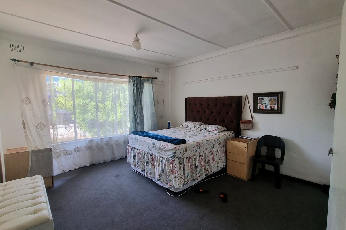 A spacious bedroom featuring large windows, carpeted flooring, and a ceiling-mounted light fixture. The room requires cosmetic updates to improve its overall condition.