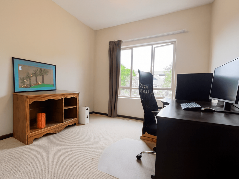 A functional bedroom or home office space featuring neutral wall tones, carpeted flooring, and a large window providing natural light.