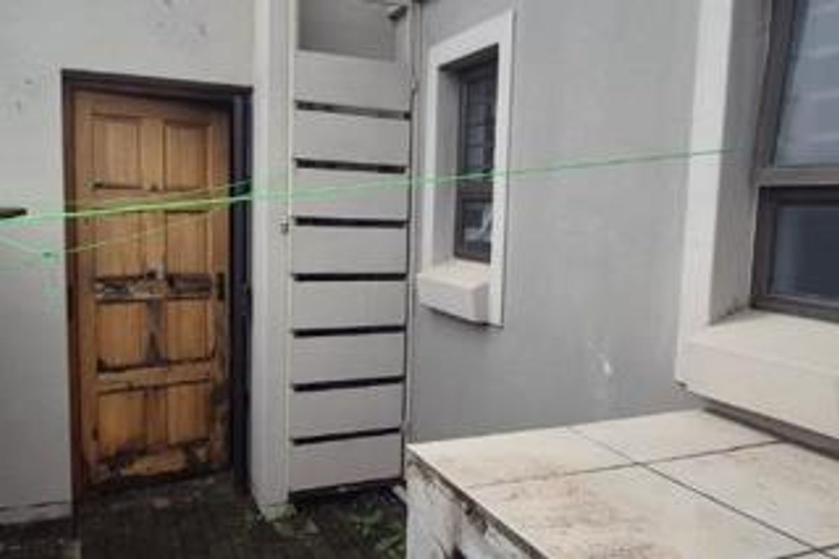 A rear exterior utility area featuring a tiled counter, security gate, and a weathered timber door requiring maintenance.