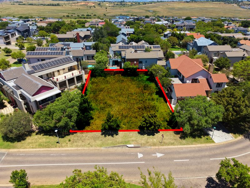 An aerial view of a vacant residential plot situated in a developed estate, featuring established trees and direct street access.