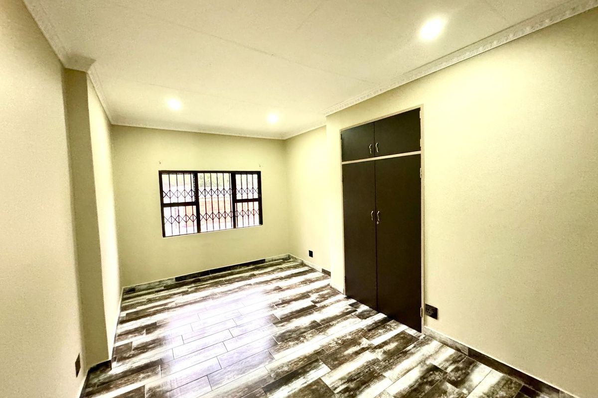 A bright, empty bedroom featuring modern wood-look tiled flooring, built-in wardrobe storage, and security bars on the window.
