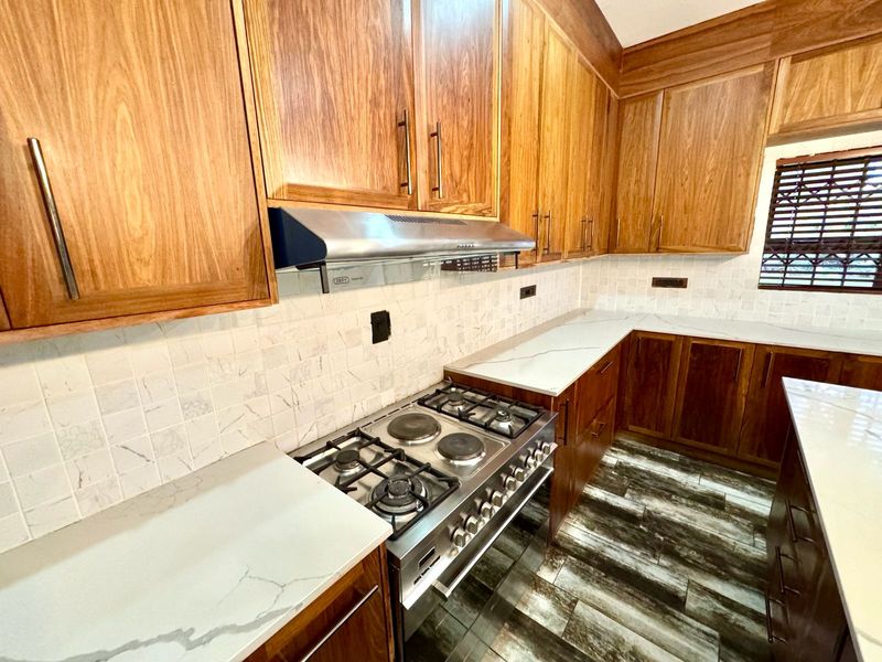 Modern kitchen featuring warm wooden cabinetry, stone countertops, a tiled backsplash, and a professional-style gas and electric hob with an integrated extractor fan.
