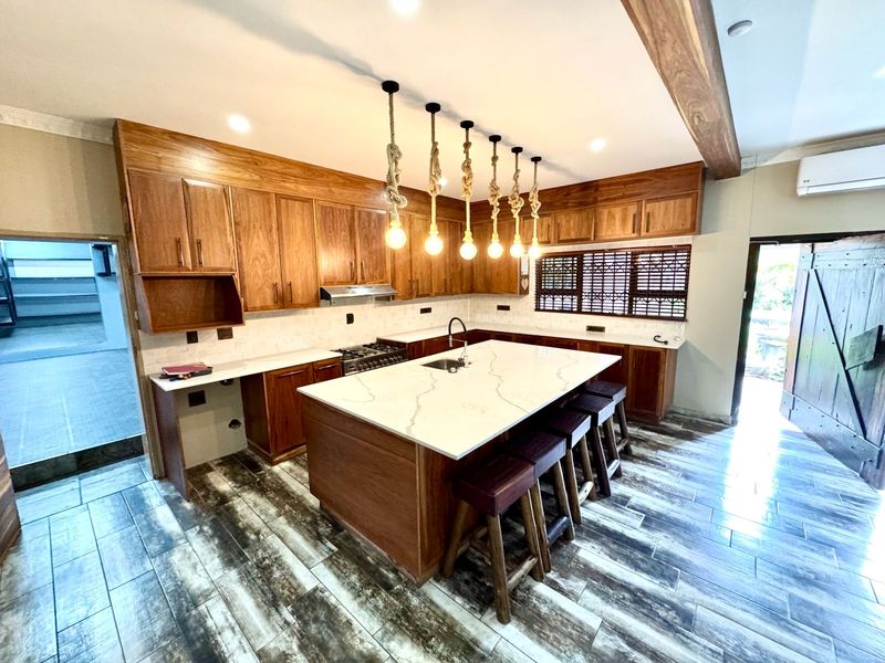 Modern kitchen featuring warm wooden cabinetry, a large stone-topped island with seating, integrated appliances, and stylish pendant lighting throughout the space.