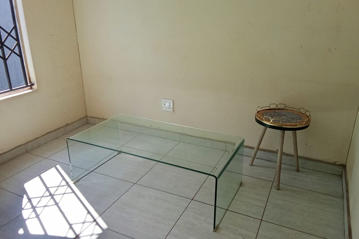 A sparsely furnished living area featuring tiled flooring, a window with a security gate, and neutral wall finishes.