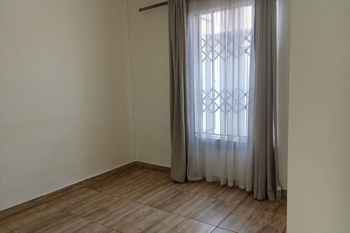 A clean, empty bedroom featuring wood-look tiled flooring and a window fitted with a security gate and curtain rod.