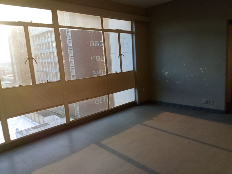 An empty living area featuring large metal-framed windows and tiled flooring, requiring cosmetic updates to the walls and finishes.