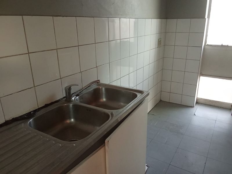 A functional kitchen area featuring a double bowl stainless steel sink, tiled splashback, and tiled flooring, currently in need of cosmetic updates.