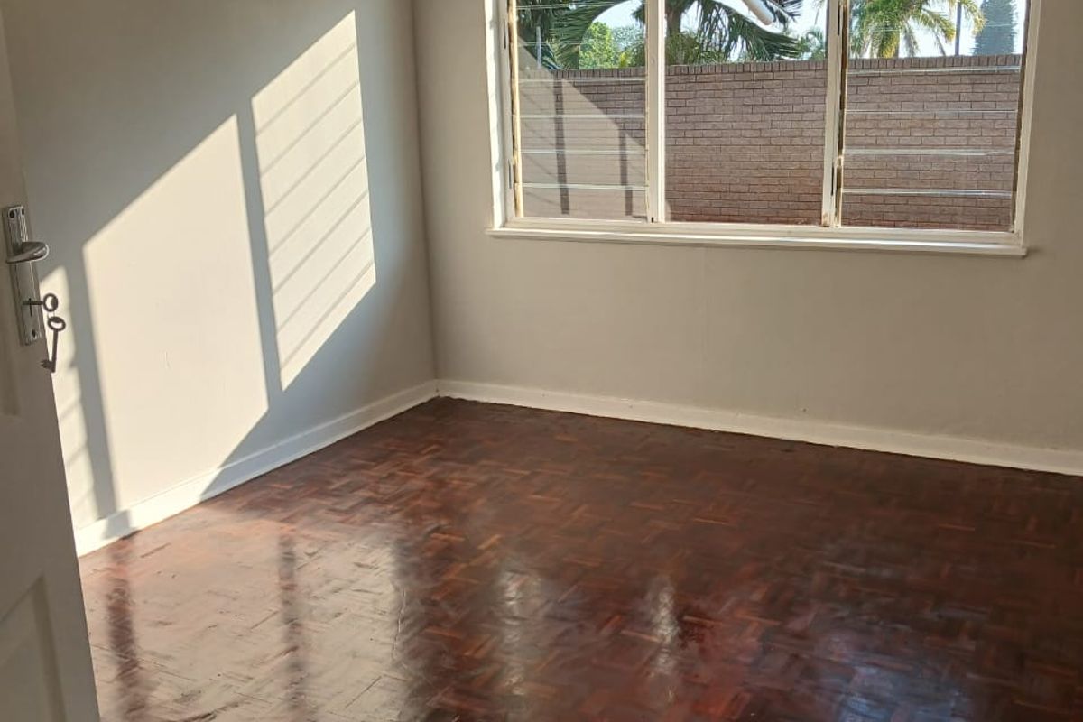 A bright, empty bedroom featuring polished parquet flooring and a large window providing natural light.