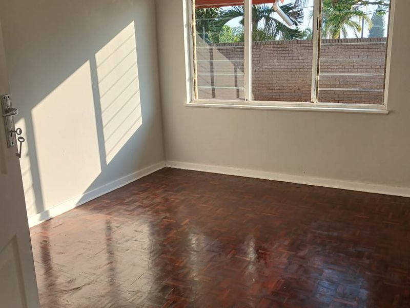 A bright, empty bedroom featuring polished parquet flooring and a large window providing natural light.