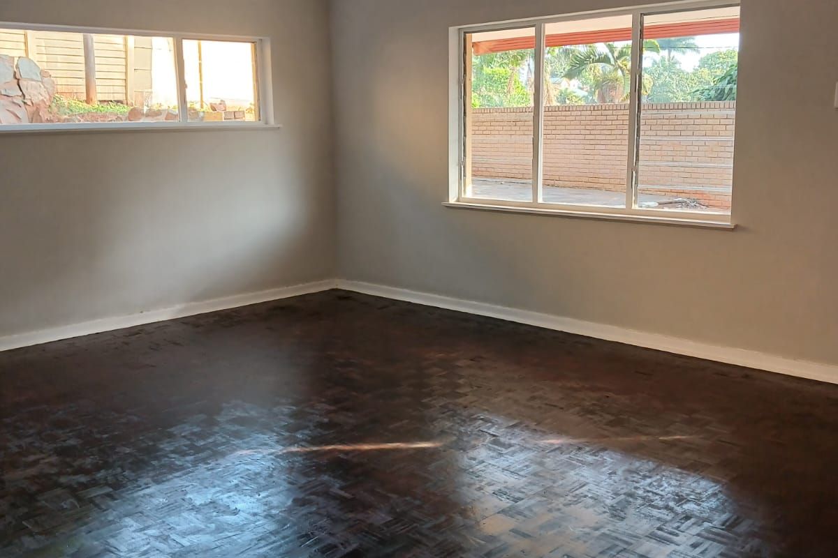 A bright, empty living room featuring dark parquet flooring, neutral walls, and large windows providing natural light.
