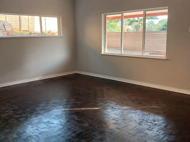 A bright, empty living room featuring dark parquet flooring, neutral walls, and large windows providing natural light.