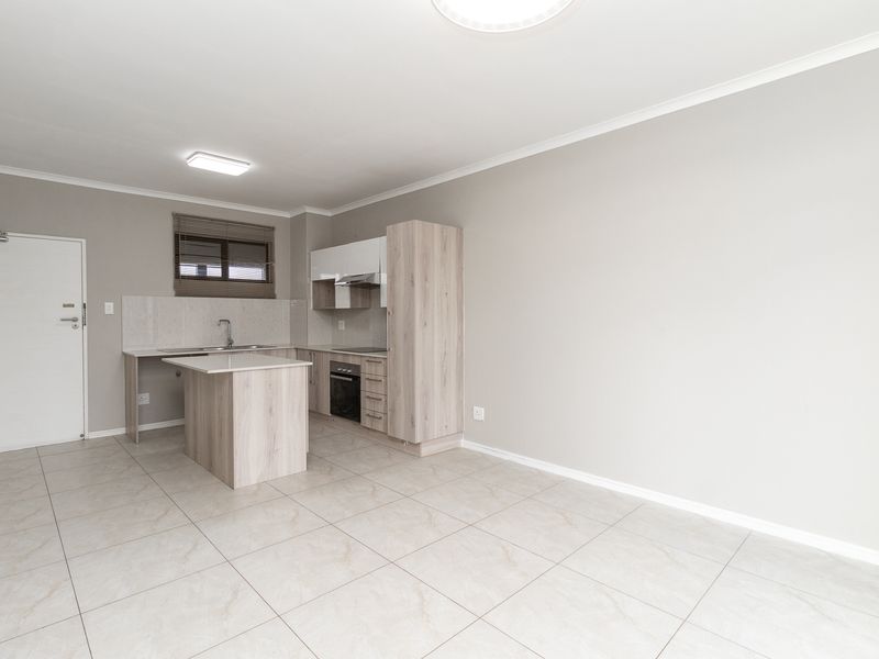 Modern open-plan kitchen and living area featuring light wood-finish cabinetry, integrated appliances, and neutral tiled flooring in a clean, vacant space.