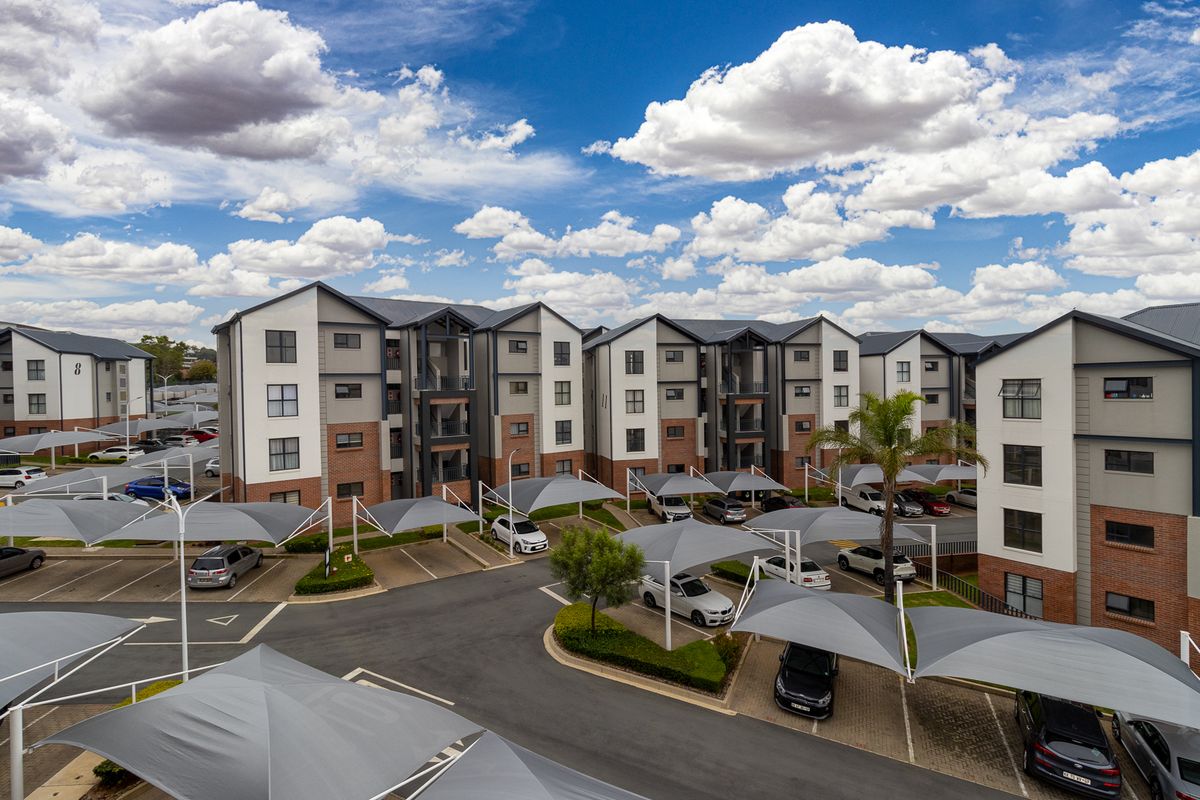 Aerial view of a modern apartment complex featuring multi-story residential buildings, paved parking areas with shade cloth carports, and landscaped grounds.